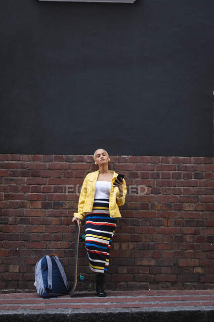 Смешанная расовая альтернатива женщина с короткими светлыми волосами в городе, опираясь на стену, используя смартфон с беспроводными наушниками и держа скейтборд. Городской цифровой кочевник на ходу. — стоковое фото
