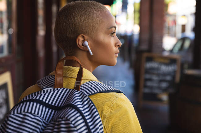 Mixed Race Alternative Frau mit kurzen blonden Haaren an einem sonnigen Tag in der Stadt unterwegs, trägt kabellose Kopfhörer und einen Rucksack, geht und schaut weg. Urbaner digitaler Nomade unterwegs. — Stockfoto