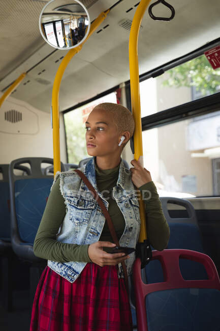 Femme alternative de race mixte avec les cheveux blonds courts sur et autour dans la ville, debout sur un bus en utilisant un smartphone avec écouteurs sans fil et détournant les yeux. Nomade numérique urbain en déplacement. — Photo de stock