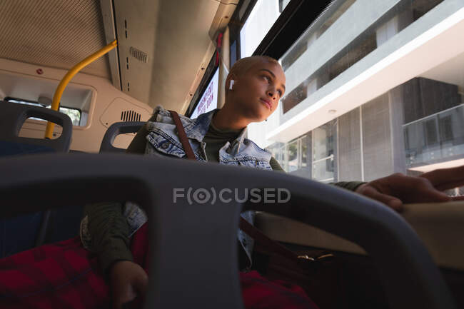 Razza mista donna alternativa con i capelli corti biondi in giro per la città, seduto su un autobus con gli auricolari wireless e guardando fuori dalla finestra. Nomade digitale urbano in movimento. — Foto stock