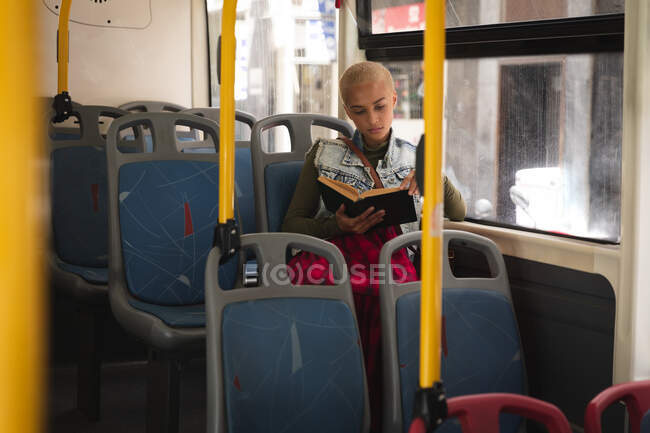 Mujer alternativa de raza mixta con pelo corto y rubio en la ciudad, sentada en un autobús leyendo un libro. Nómada urbano independiente en movimiento. - foto de stock