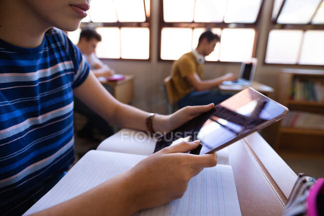 Вид збоку посередині секції змішаної раси школярка-підліток, що сидить за столом у класі, використовуючи планшетний комп'ютер, з однокласниками, що працюють на задньому плані — стокове фото