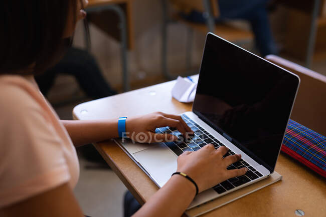 Vista lateral sección media de una adolescente de raza mixta sentada en un escritorio en clase usando una computadora portátil - foto de stock