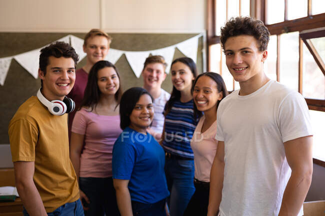 Vista frontal de un grupo multiétnico de alumnos adolescentes de la escuela de pie juntos en un aula y sonriendo a la cámara en el momento del descanso - foto de stock