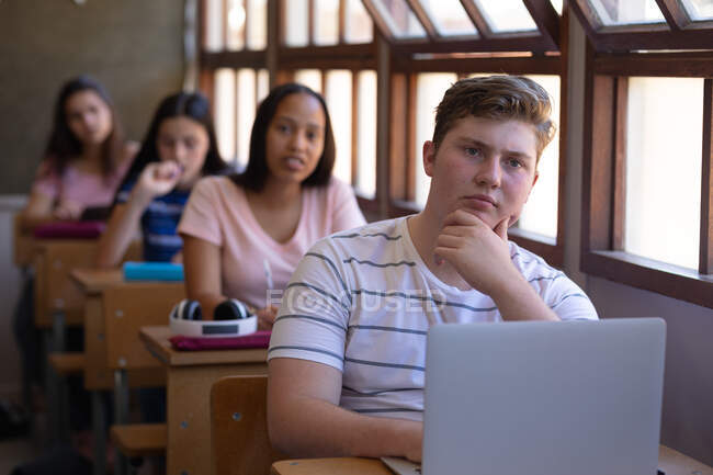 На перший погляд на підлітка, який сидить за столом і дивиться вперед, і зосереджується в класі, з рядом однокласниць-підлітків, які сидять за партами позаду нього. — стокове фото