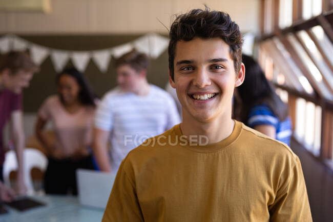Porträt eines kaukasischen Teenagers mit kurzen dunklen Haaren und grauen Augen, der in einem Klassenzimmer steht und in die Kamera lächelt, während sich Mitschüler im Hintergrund unterhalten. — Stockfoto