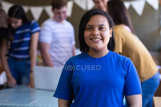 Portrait close up of a mixed race teenage girl with dark hair and brown eyes standing in a school classroom smiling to camera, with classmates talking in the background — Stock Photo