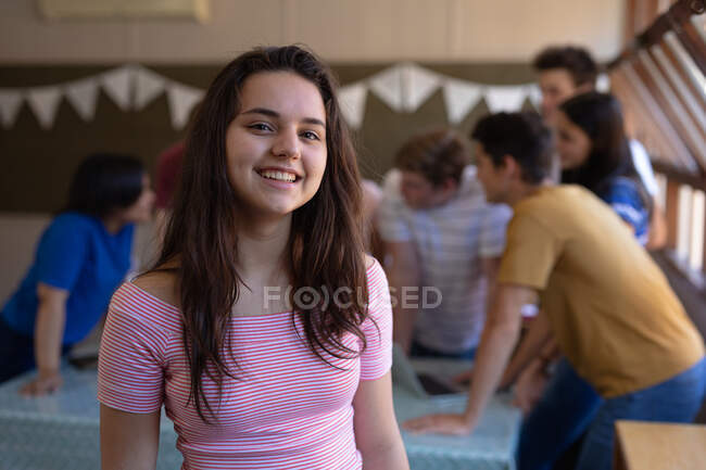Retrato de una adolescente caucásica con el pelo largo y oscuro y los ojos marrones de pie en un aula de la escuela sonriendo a la cámara, con compañeros de clase hablando en el fondo - foto de stock