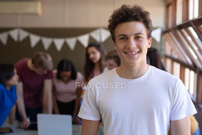 Retrato de cerca de un adolescente caucásico con el pelo corto y oscuro y los ojos grises de pie en un aula de la escuela sonriendo a la cámara, con compañeros de clase hablando en el fondo - foto de stock
