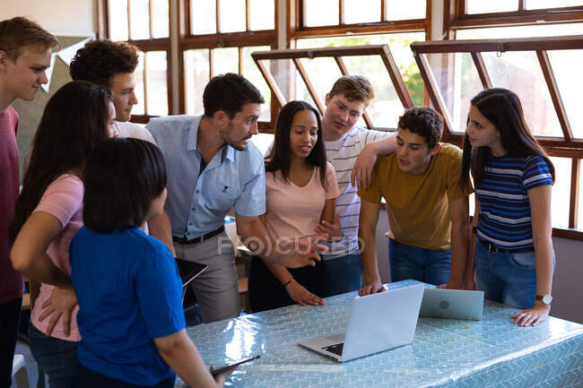 Vista frontal de um grupo multi-étnico de alunos da escola adolescente e seu professor masculino caucasiano em pé em uma sala de aula olhando para computadores portáteis juntos, os alunos ouvindo enquanto o professor fala — Fotografia de Stock