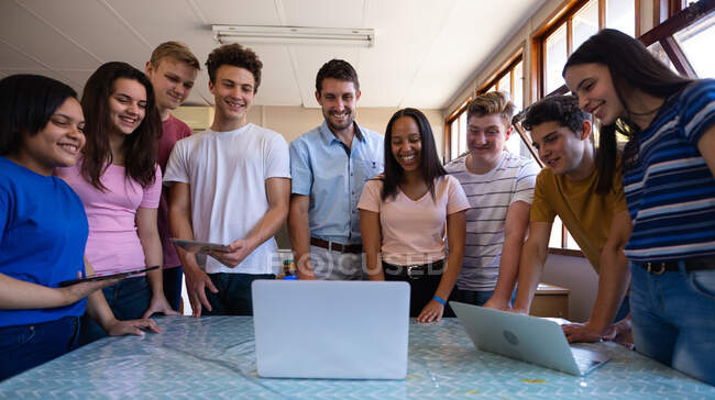 Vista frontale di un gruppo multietnico di alunni adolescenti e del loro insegnante maschio caucasico in piedi in un'aula a guardare computer portatili insieme e sorridere — Foto stock