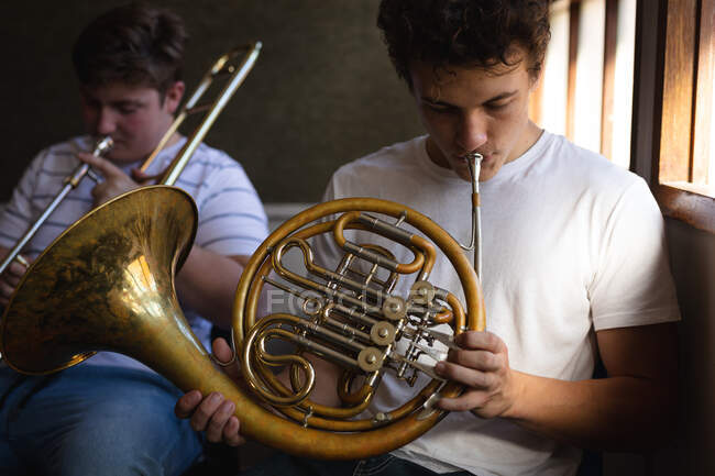 Vue de face de deux musiciens masculins adolescents caucasiens qui répètent, l'un jouant du cor français et l'autre du trombone. Concept d'enseignement secondaire. — Photo de stock