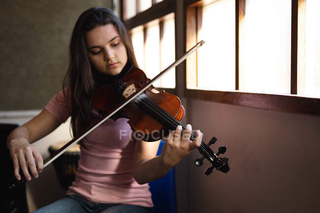 Портрет кавказького музиканта, гарної дівчини підліткового віку, що сидить біля вікна і грає на скрипці у старшій школі. — стокове фото