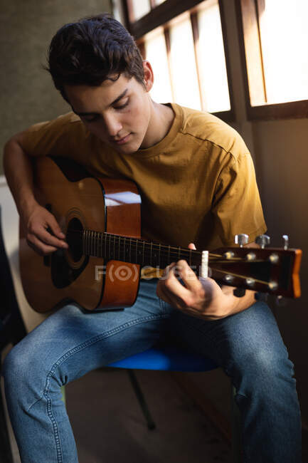 Vista frontale da vicino di un ragazzo adolescente musicista caucasico seduto e che suona una chitarra acustica in una scuola superiore — Foto stock