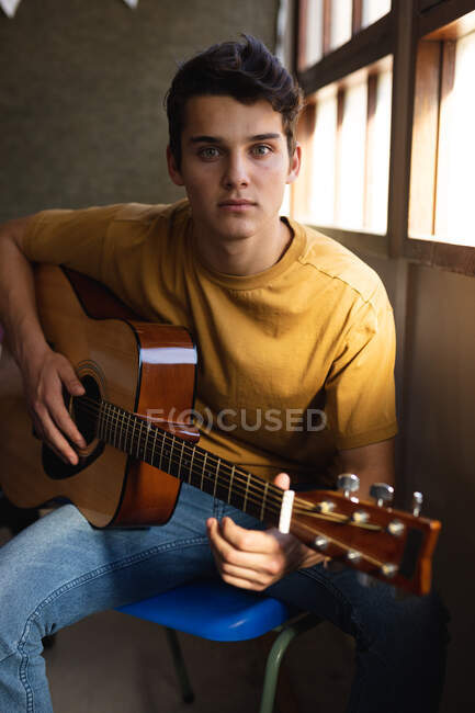Retrato de perto de um adolescente caucasiano sentado à janela segurando uma guitarra acústica e olhando para a câmera em uma escola — Fotografia de Stock