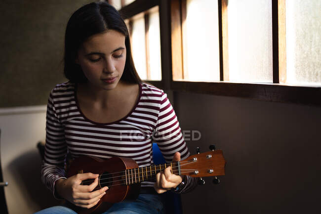 Porträt eines kaukasischen Musikers Teenager-Mädchen, das am Fenster sitzt, nach unten schaut und in einer High School Ukulele spielt — Stockfoto