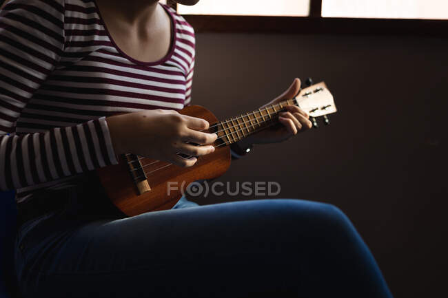 Vista lateral sección media de músico adolescente sentado junto a una ventana en una sombra y tocando un ukelele - foto de stock