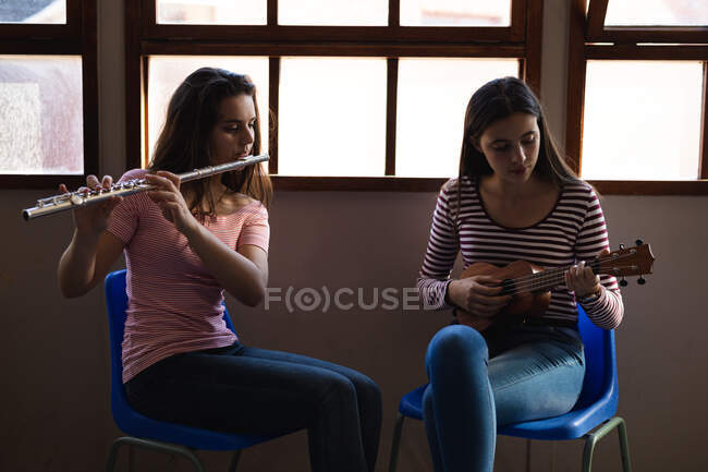 Vorderansicht zweier kaukasischer Teenager mit langen dunklen Haaren, die vor einem Fenster sitzen und Flöte und Ukulele spielen — Stockfoto