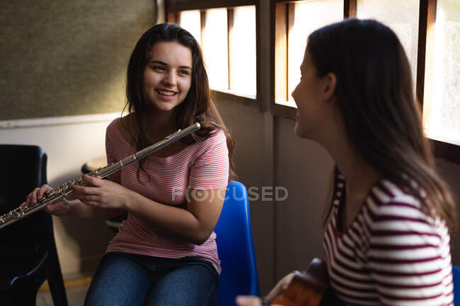 Vista lateral de dos adolescentes caucásicas con el pelo largo y oscuro sentadas frente a una ventana sosteniendo una flauta y un ukelele mirándose y sonriendo - foto de stock