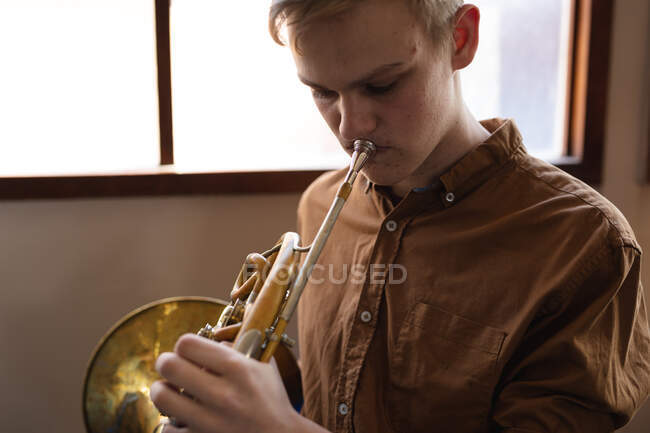 Vista frontale primo piano di un ragazzo caucasico musicista adolescente con camicia marrone seduto davanti a una finestra che suona un corno francese da solo a scuola — Foto stock
