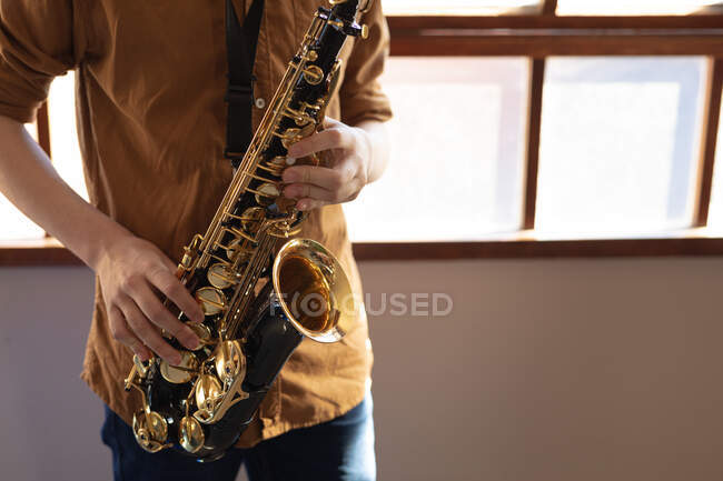 Vista frontal sección media del músico adolescente de pie tocando un saxofón frente a una ventana durante una práctica de banda escolar - foto de stock