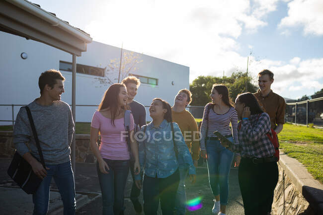 Vista frontal de un grupo multiétnico de estudiantes adolescentes masculinos y femeninos que hablan mientras caminan por sus terrenos escolares - foto de stock