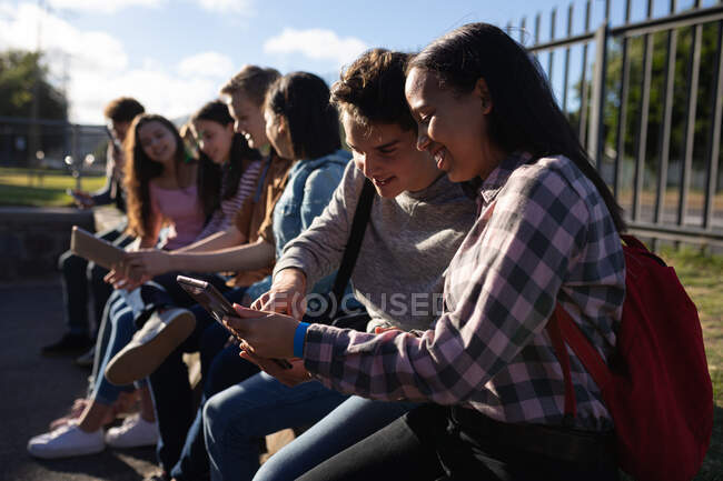 Вид сбоку на многоэтническую группу студентов-подростков, сидящих на стене и говорящих за компьютерами на территории школы — стоковое фото