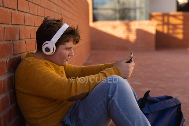 Vista lateral de cerca de un adolescente caucásico sentado solo en un patio de la escuela con auriculares y mirando un teléfono inteligente - foto de stock