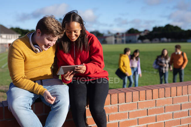 Vue de face d'un adolescent caucasien et d'une adolescente métisse assise sur un mur regardant ensemble une tablette et souriant, dans un terrain de jeu d'école avec deux adolescents marchant en arrière-plan — Photo de stock