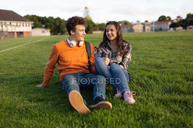 Передній вид Кавказької дівчини - підлітка і хлопчика посміхаються один до одного і тримаються за руки, сидячи в школі, граючи на полі. — стокове фото