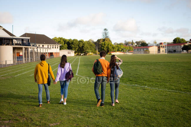 Vista trasera de dos parejas adolescentes caucásicas con bolsas de escuela cogidas de la mano y caminando a través de un campo de juego de la escuela en un día soleado - foto de stock