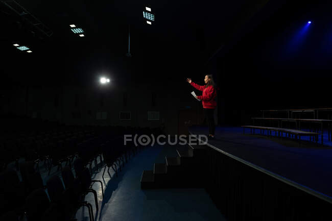 Vista laterale di un'adolescente mista in piedi sul palco con una sceneggiatura in un teatro scolastico vuoto durante le prove per uno spettacolo — Foto stock