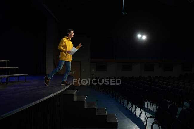 Seitenansicht eines kaukasischen Teenagers, der während der Proben für eine Aufführung in einem leeren Schultheater auf der Bühne steht und ein Drehbuch hält — Stockfoto
