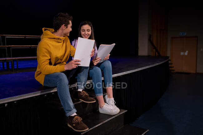 Vista frontal de una adolescente y un niño caucásicos sentados en el borde del escenario sosteniendo guiones y sonriéndose en un teatro escolar vacío durante los ensayos para una actuación - foto de stock