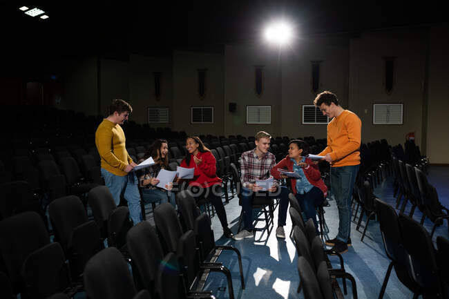 Frontansicht einer multiethnischen Gruppe von Teenagern im Gespräch, Drehbücher haltend und lächelnd in der Aula eines leeren Schultheaters während der Proben für eine Aufführung — Stockfoto