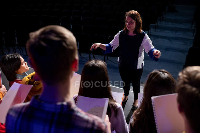Over the shoulder view of a Caucasian female conductor and a multi-ethnic group of teenage male and female choristers standing opposite each other, the choir holding sheet music and singing — Stock Photo
