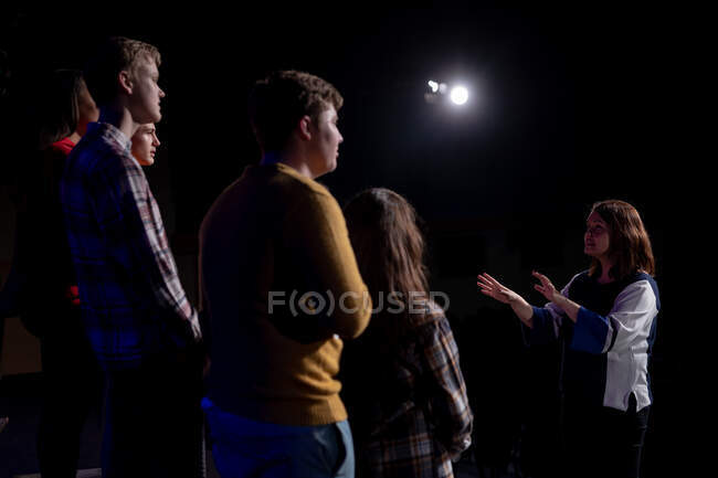 Vista lateral de una directora de orquesta caucásica y un grupo multiétnico de coristas adolescentes masculinos y femeninos de pie enfrente, cantando en el escenario de un teatro escolar durante los ensayos para una actuación - foto de stock