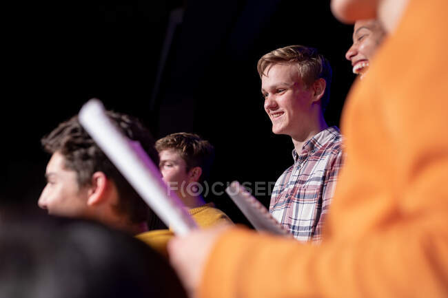 Vista lateral de cerca de un grupo multiétnico de coristas adolescentes masculinos y femeninos sosteniendo partituras y riendo, de pie en el escenario de un teatro escolar durante una pausa en los ensayos para una actuación - foto de stock