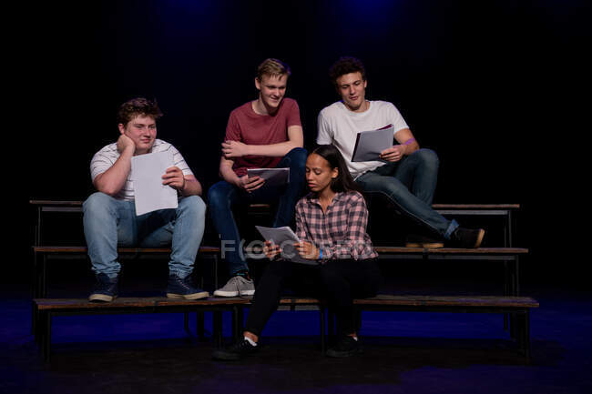Vue de face de trois adolescents caucasiens et d'une adolescente métisse tenant des scénarios et souriant, assis sur la scène d'un théâtre scolaire pendant les répétitions pour une performance — Photo de stock