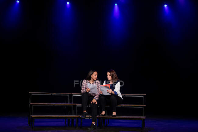 Передній погляд на кавказьку і змішану расу дівчаток-підлітків, що ведуть сценарії сидячі на сцені шкільного театру під час репетицій для виступу. — стокове фото