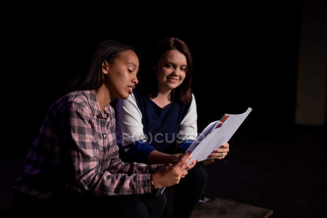 Побочный вид кавказки и смешанной расы девочки-подростки, держащие сценарии, сидящие на сцене школьного театра во время репетиций спектакля — стоковое фото