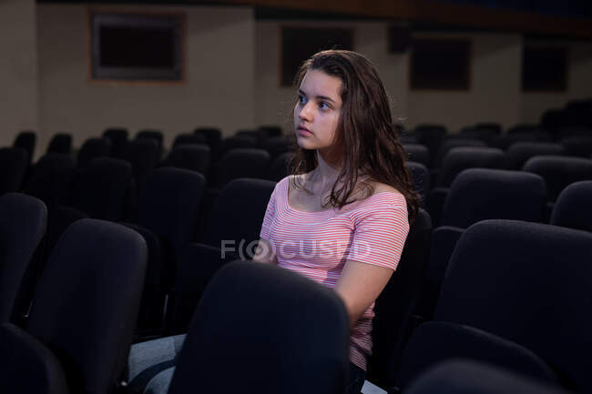 Vista frontal de una adolescente caucásica sentada en el auditorio vacío de un teatro escolar mirando a sus amigos en el escenario durante los ensayos para una actuación - foto de stock