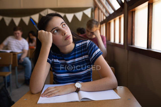 Вид спереди на кавказскую девочку-подростка, сидящую за столом в школьном классе, смотрящую в окно, с одноклассниками, сидящими за партами на заднем плане — стоковое фото