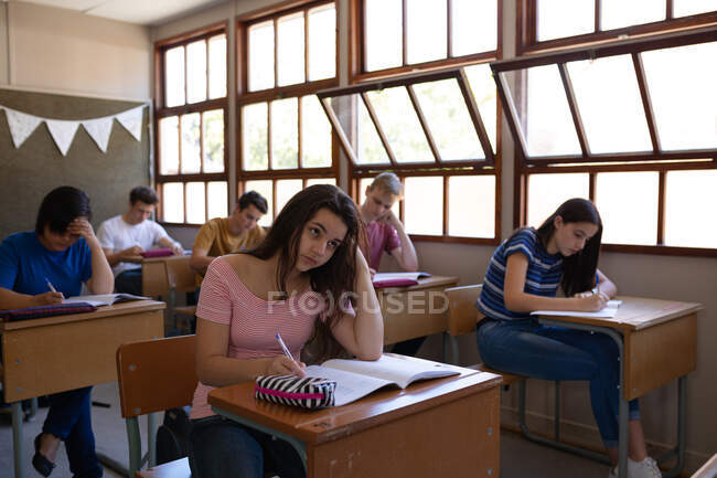 Vista frontal de un grupo multiétnico de alumnos adolescentes que se concentran sentados en escritorios en clase estudiando en una escuela secundaria - foto de stock