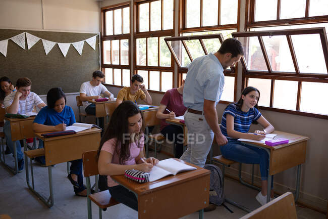 Vista lateral de un grupo multiétnico de alumnos adolescentes sentados en escritorios en clase estudiando en la escuela con un profesor caucásico de pie y hablando con una chica caucásica en su escritorio - foto de stock