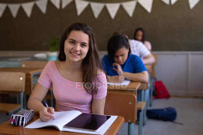 Portrait of a Caucasian teenage girl sitting at a desk in a school classroom writing in a notebook and looking to camera smiling, with classmates sitting at desks working in the background — Stock Photo