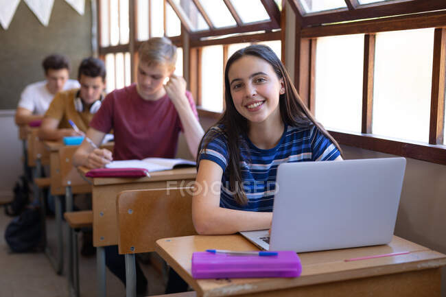 Retrato de una adolescente caucásica sentada en un escritorio en un aula de la escuela usando una computadora portátil y mirando a la cámara sonriendo, con compañeros de clase sentados en escritorios trabajando en el fondo - foto de stock