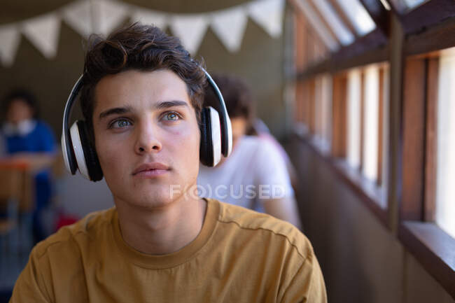 Retrato de perto de um adolescente caucasiano com cabelos escuros e olhos grisalhos sentado em uma mesa em uma sala de aula da escola usando fones de ouvido e olhando para uma janela — Fotografia de Stock