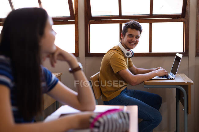 Seitenansicht eines kaukasischen Teenagermädchens, das an einem Schreibtisch in einer Schulklasse sitzt, sich auf ihren Schreibtisch lehnt und einen Teenager anlächelt, der am besten Schreibtisch sitzt, einen Laptop benutzt und sie anlächelt — Stockfoto