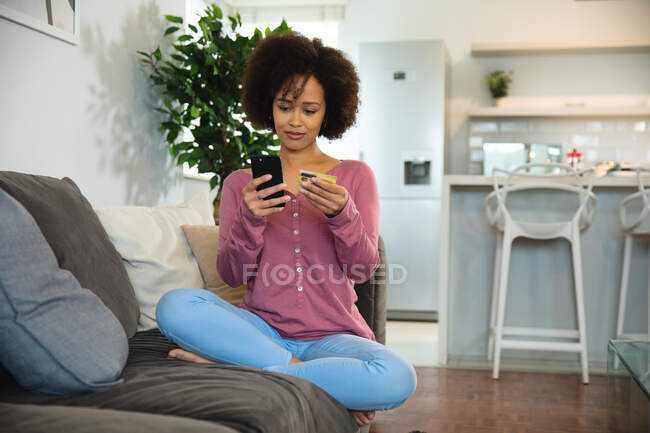 Вид спереду змішаної раси жінка розслабляється вдома, сидячи на дивані з перехрещеними ногами, тримаючи кредитну картку і використовуючи свій смартфон для здійснення онлайн транзакції — стокове фото